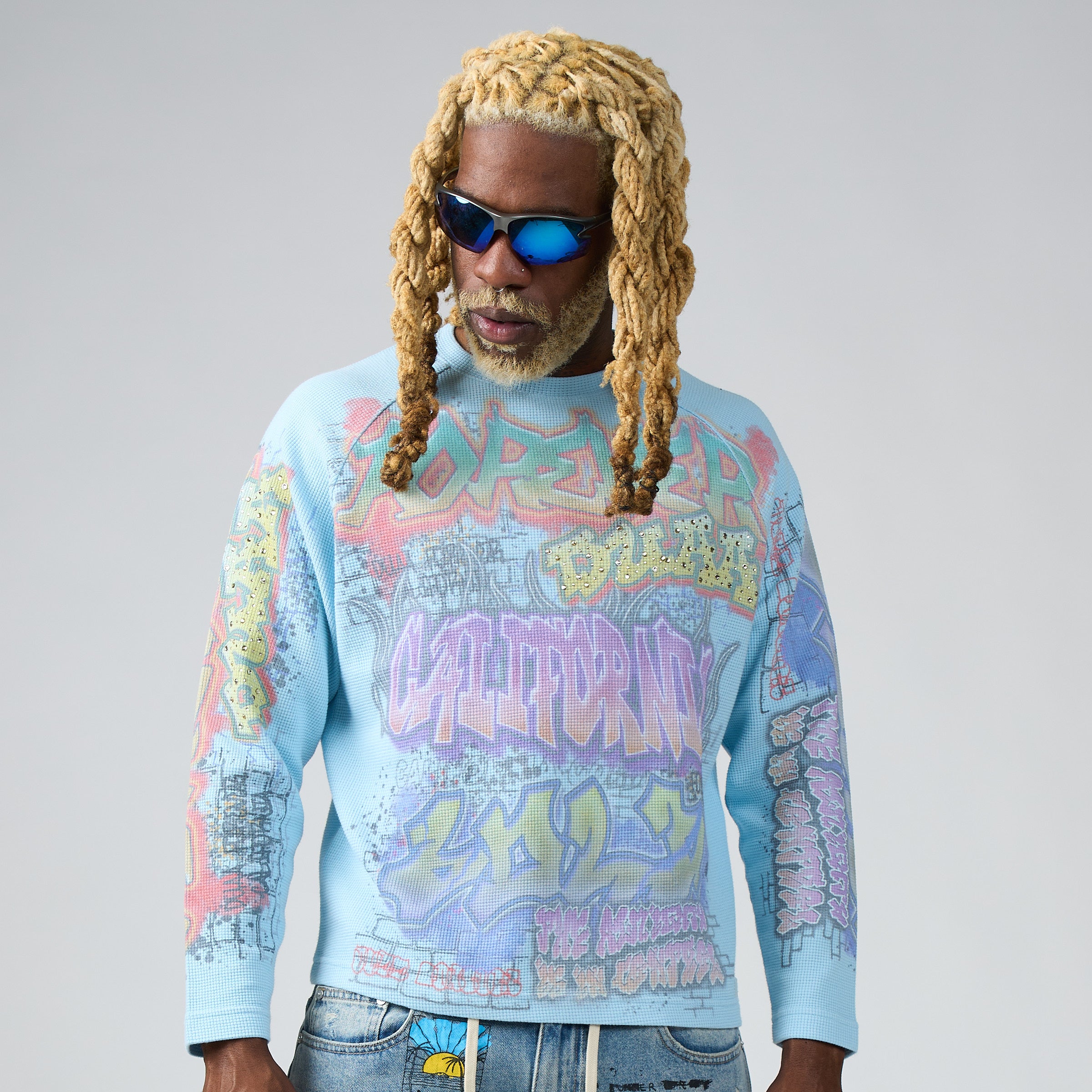 A man wearing a light blue DUAA waffle crewneck long sleeve with a colorful graffiti-style graphic print, styled with blue jeans and reflective sunglasses against a neutral gray background.