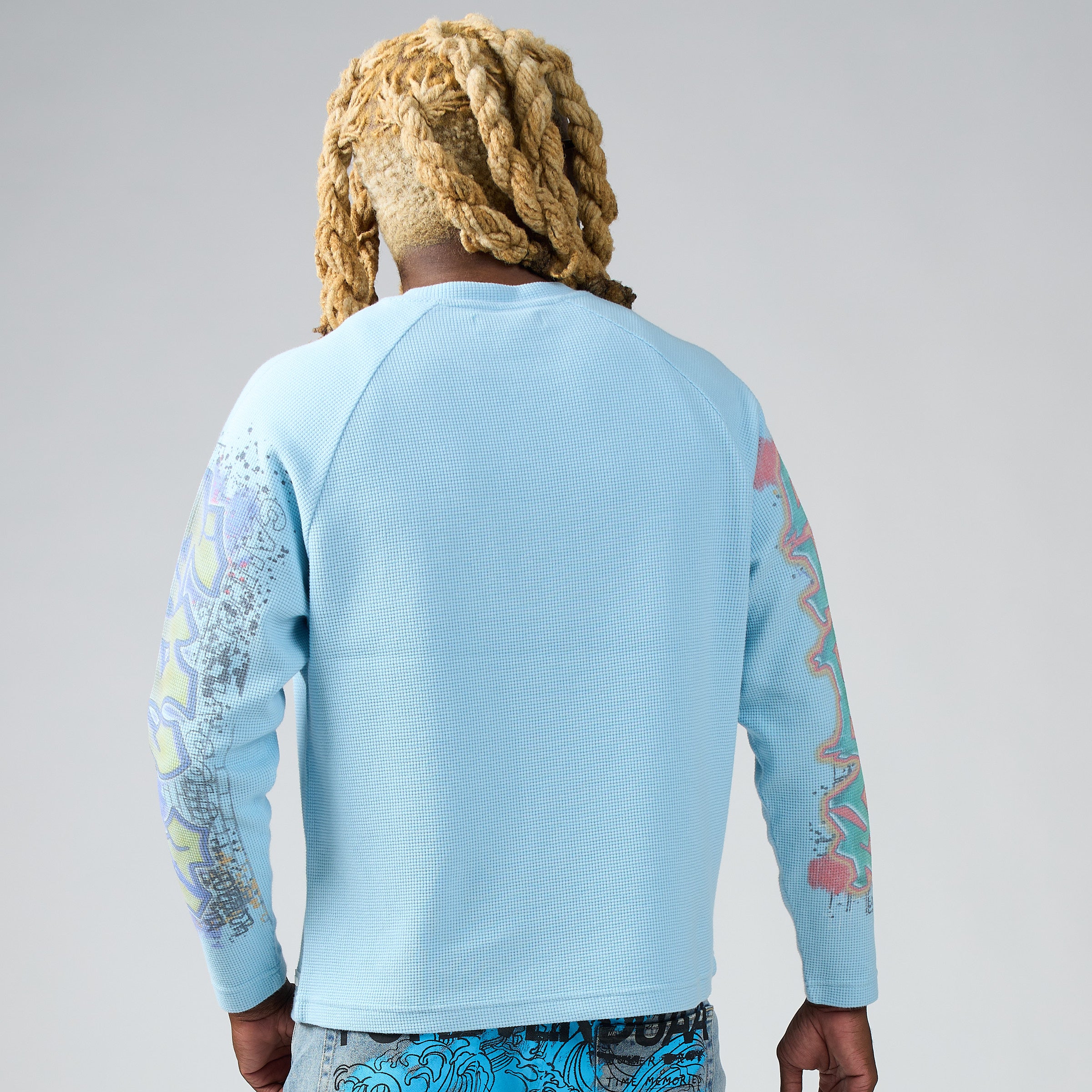 A person shown from the back wearing a light blue DUAA long-sleeve top with colorful graphic patterns on both sleeves, against a plain light gray studio background.