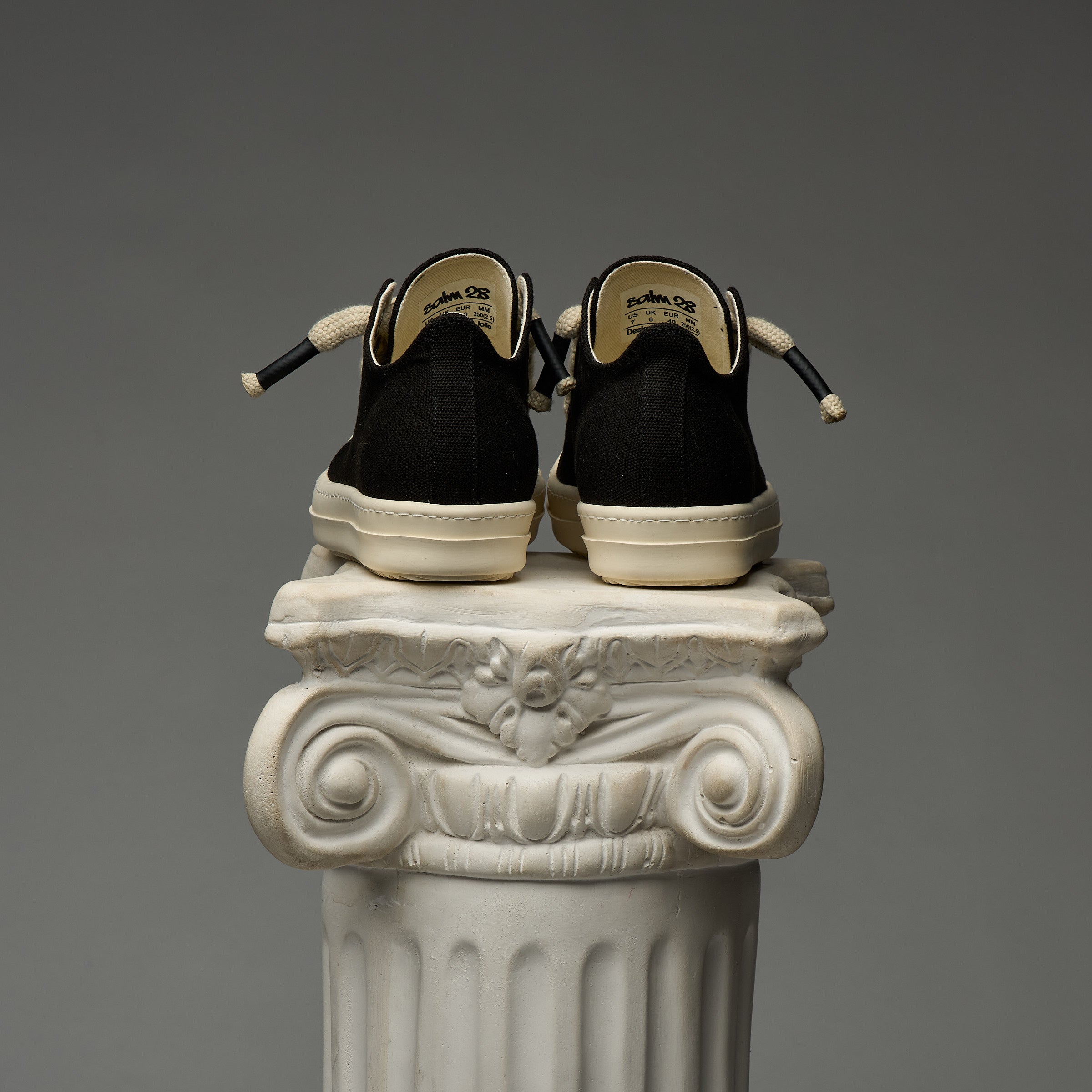 DUAA Black sneakers with white soles on a decorative column against a gray background