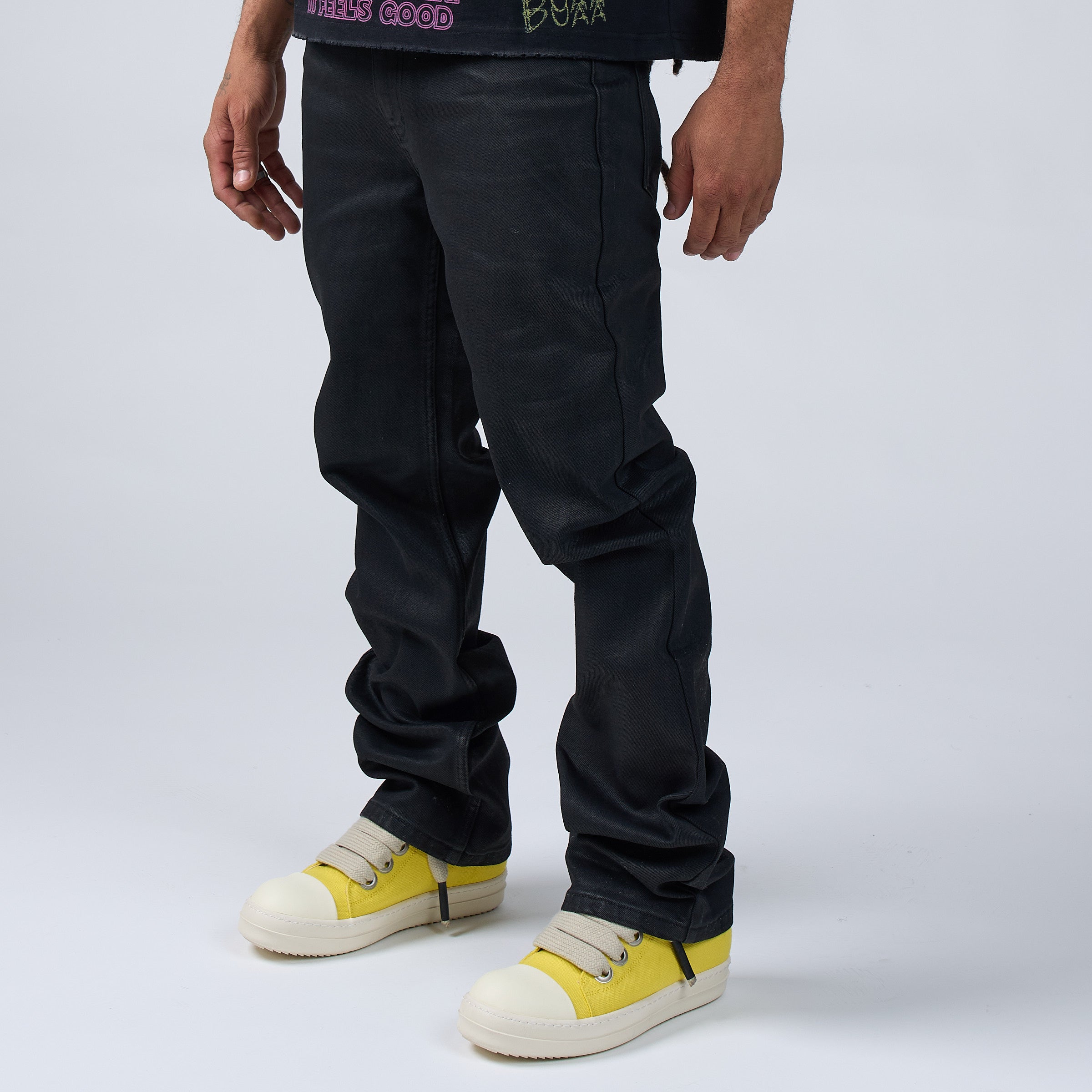 Person wearing DUAA black wax coated jeans and yellow sneakers on a light gray background