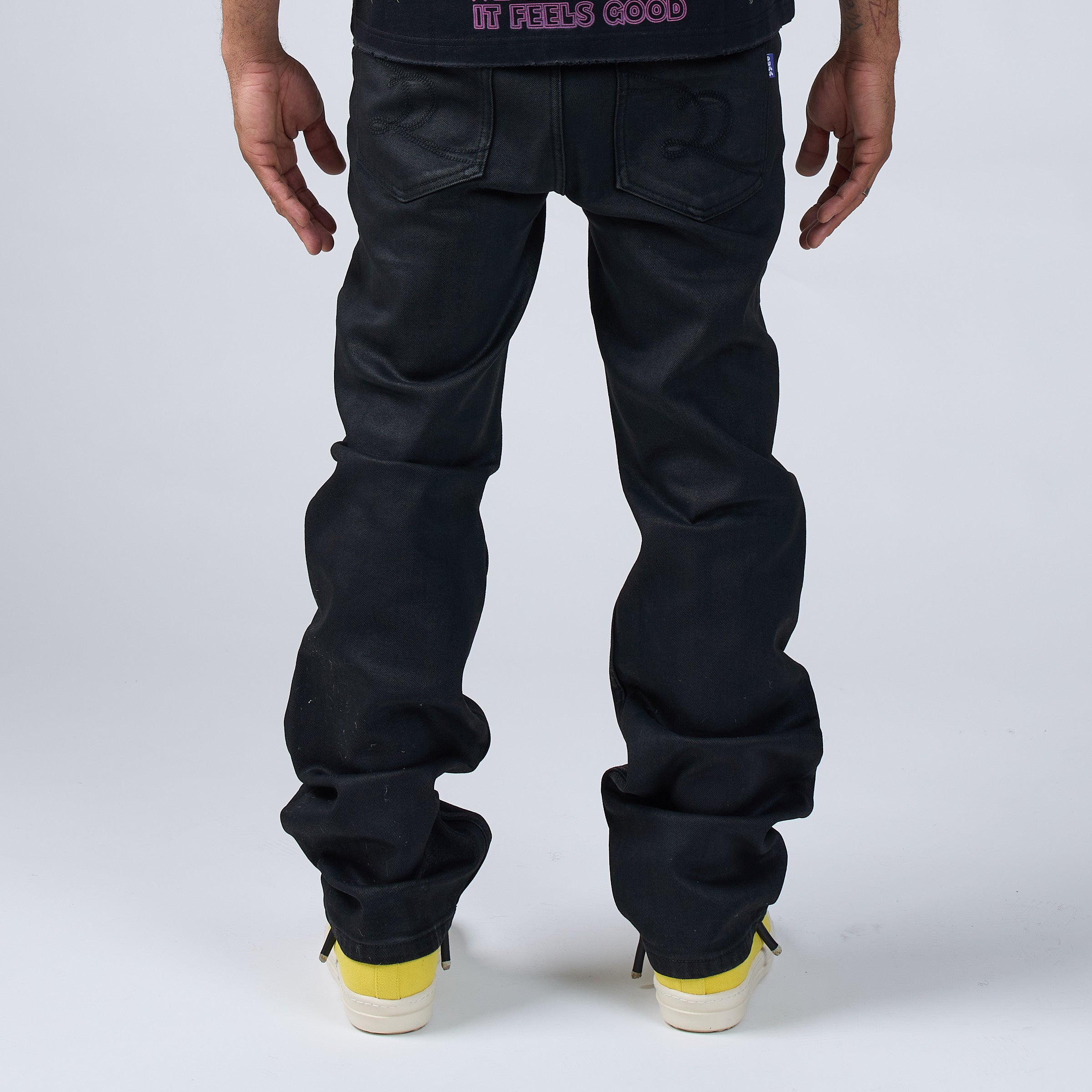 Rear view of a person wearing DUAA black wax coated jeans with cuffed ankles, paired with a dark graphic t-shirt and yellow sneakers, against a light gray studio background.