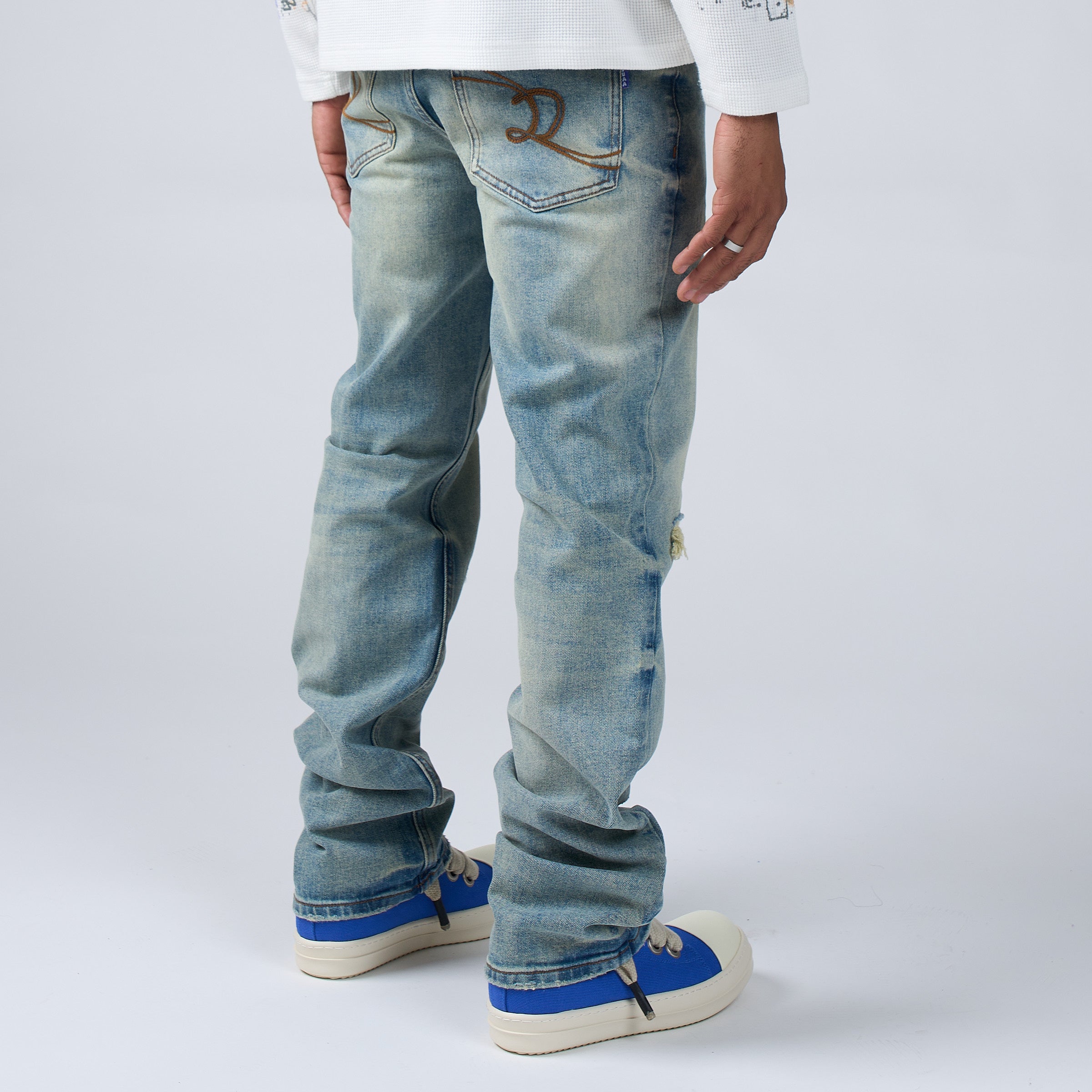 Side angle view of a Person wearing DUAA distressed blue jeans and blue sneakers on a white background