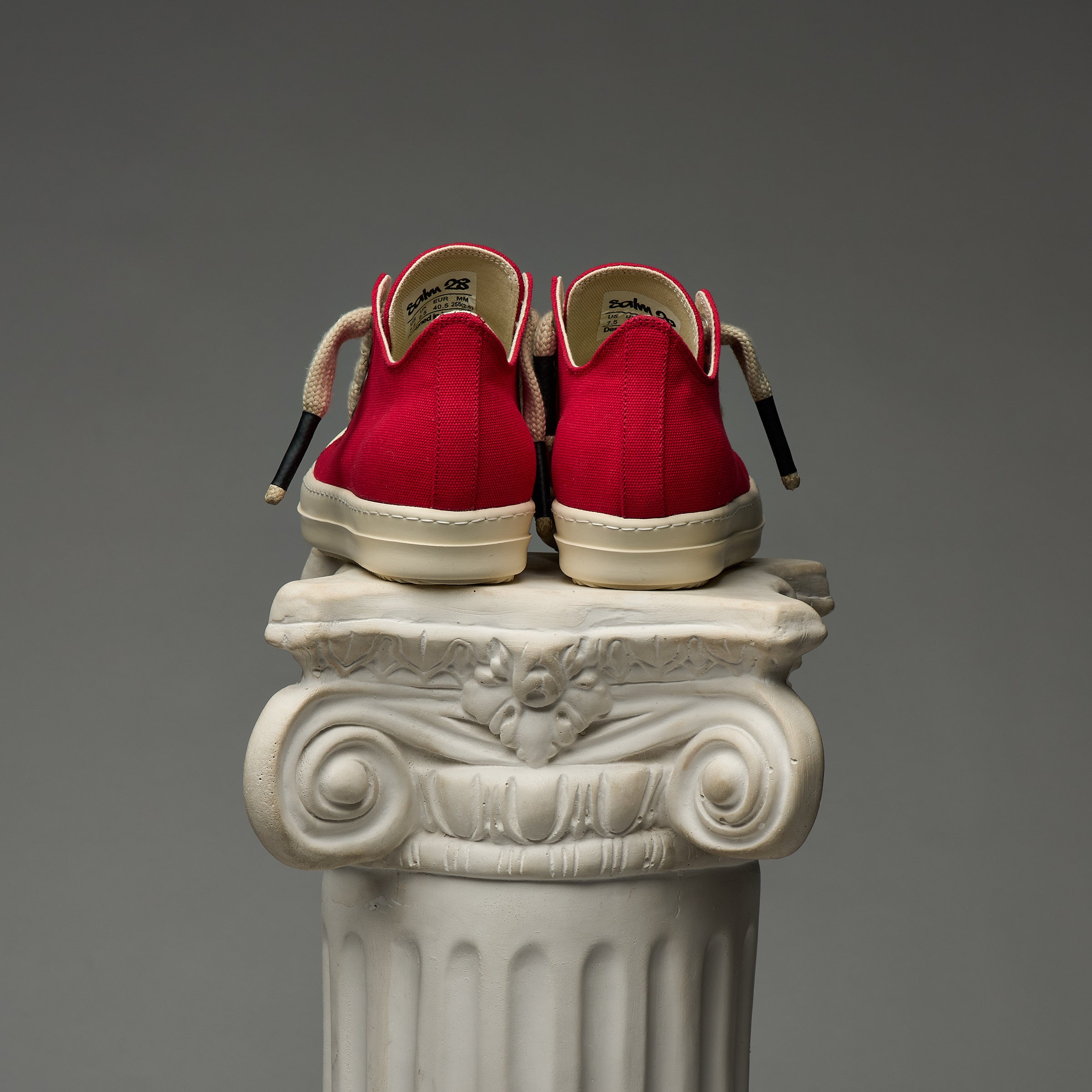 DUAA Red sneakers with white soles on a decorative column against a gray background
