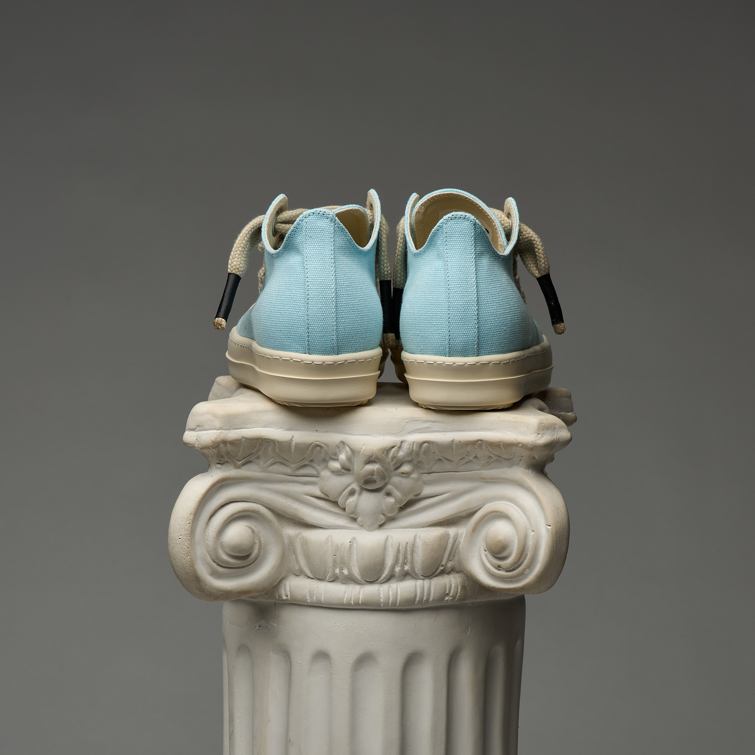 Pair of light blue DUAA sneakers with beige soles on an ornate stone column against a gray background