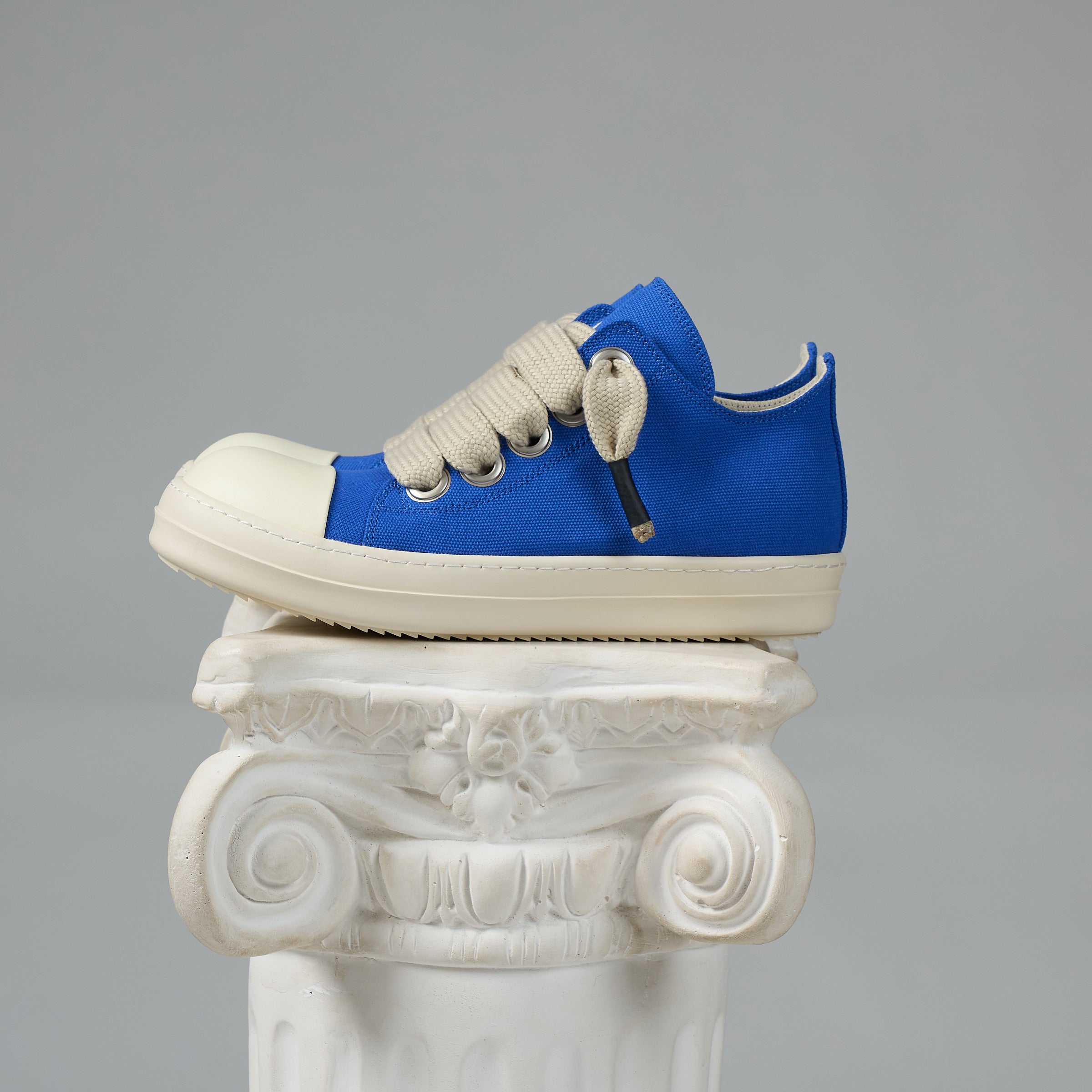 A DUAA blue canvas low-top sneaker with off-white laces and a cream rubber toe cap and sole, displayed on a white decorative column against a neutral gray background.