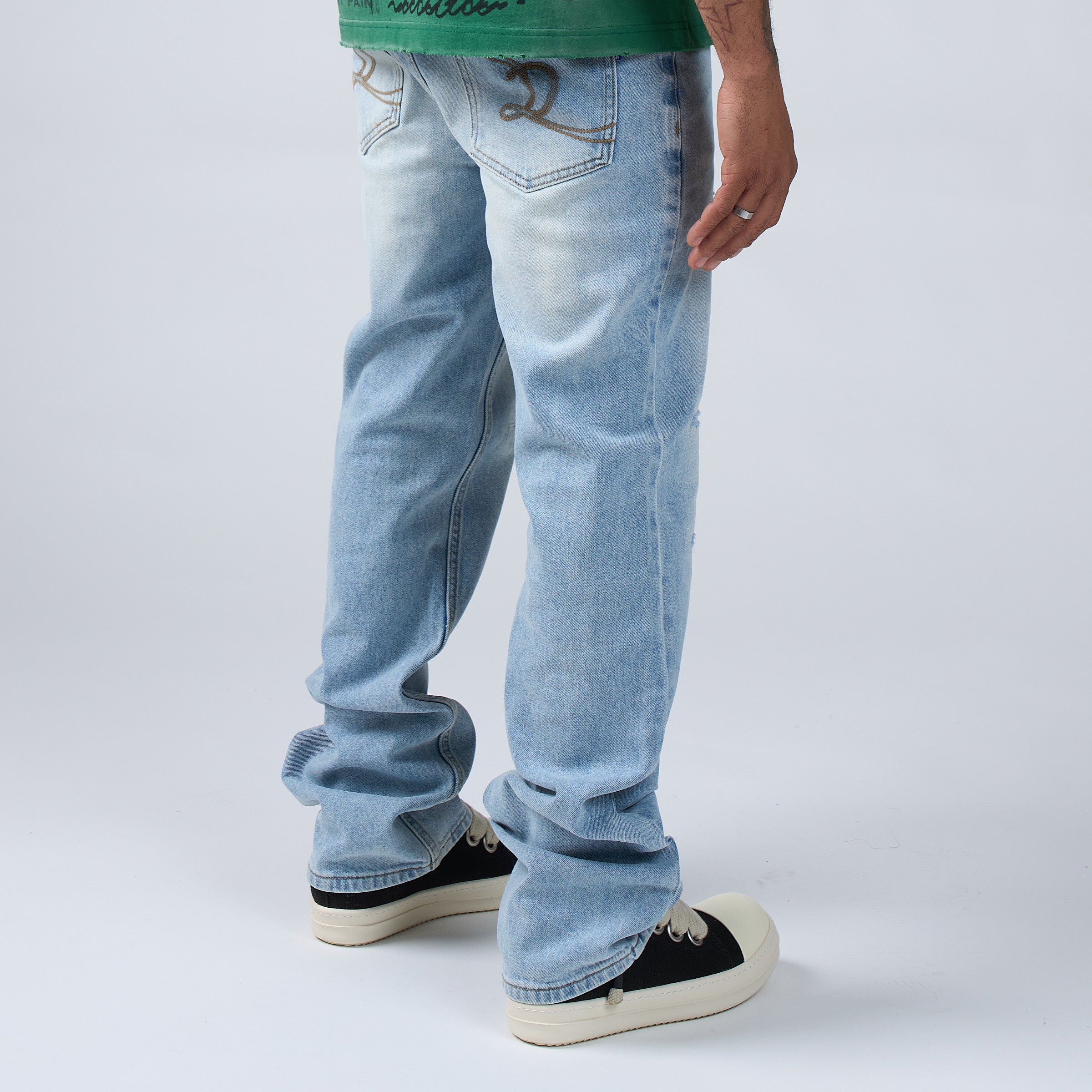 Rear side view of a person wearing DUAA ight-wash blue jeans with decorative brown embroidery on the back pockets, paired with a green graphic t-shirt and white sneakers, against a light gray studio background.