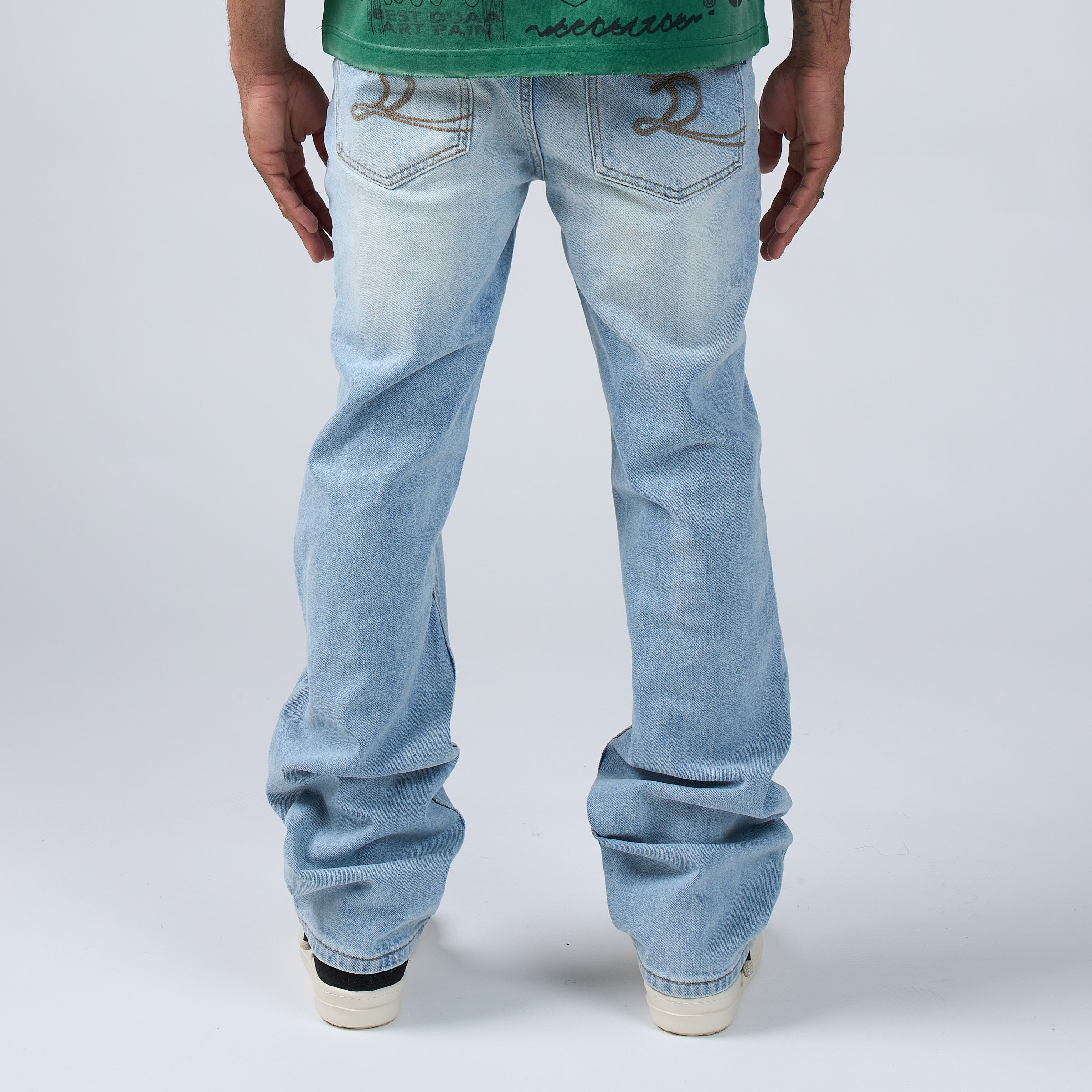 Rear view of a person wearing DUAA ight-wash blue jeans with decorative brown embroidery on the back pockets, paired with a green graphic t-shirt and white sneakers, against a light gray studio background.
