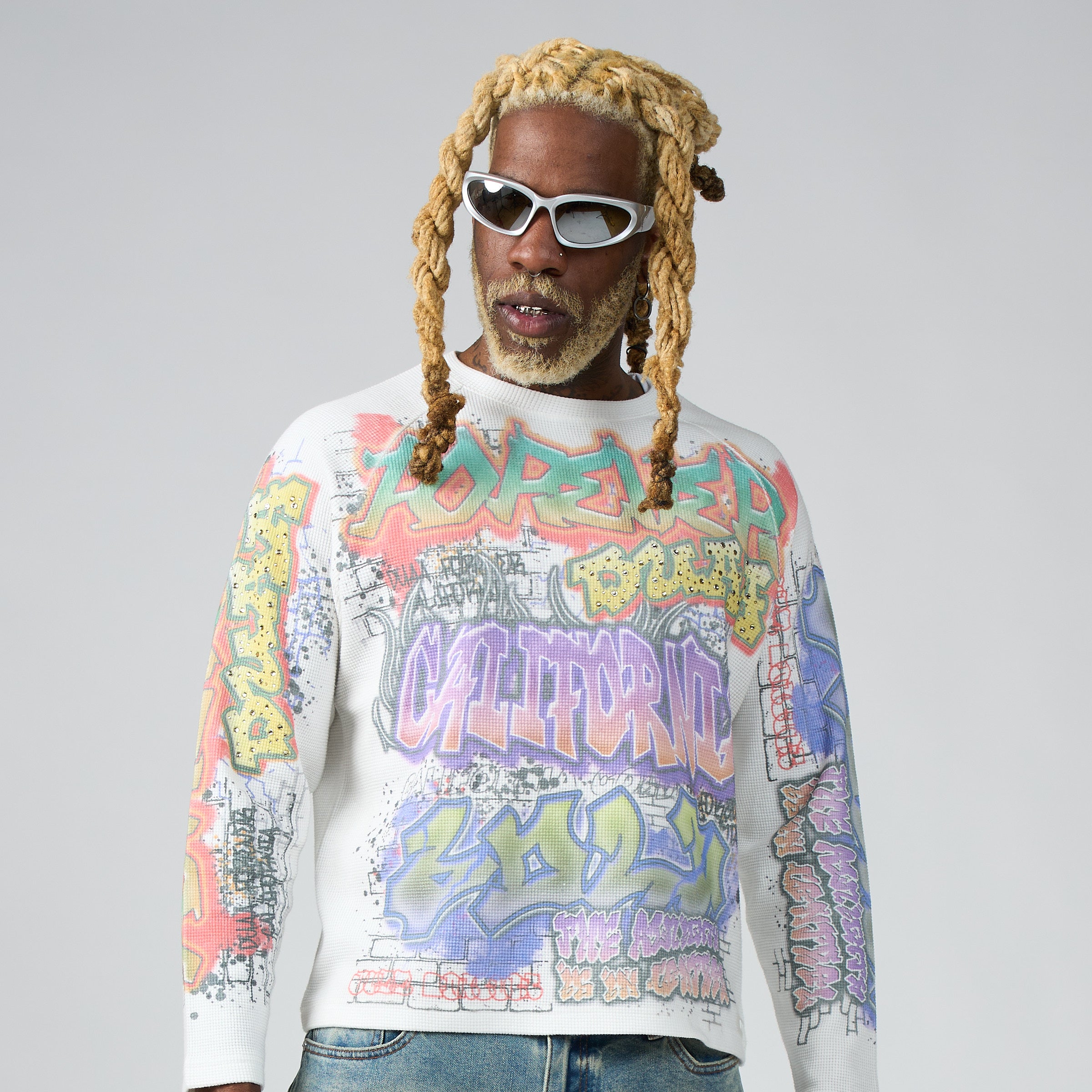 A man wearing a DUAA white Longsleeve waffle shirt with a colorful graffiti-style graphic print, styled with white sunglasses and blue jeans against a light gray studio background.