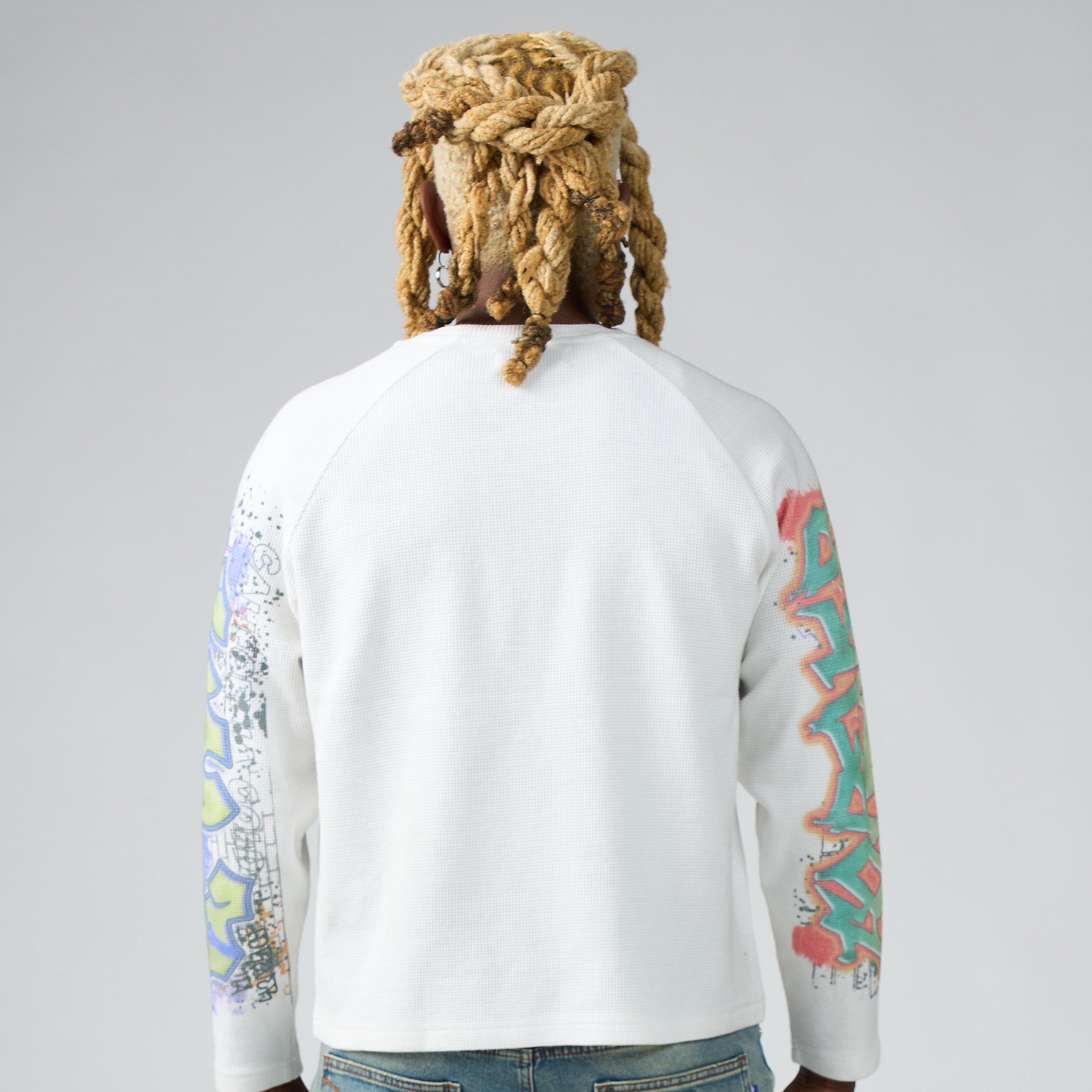 A person shown from the back wearing a DUAA white longsleeve waffle shirt with colorful graffiti-style sleeve prints, paired with blue jeans, against a light gray studio background.