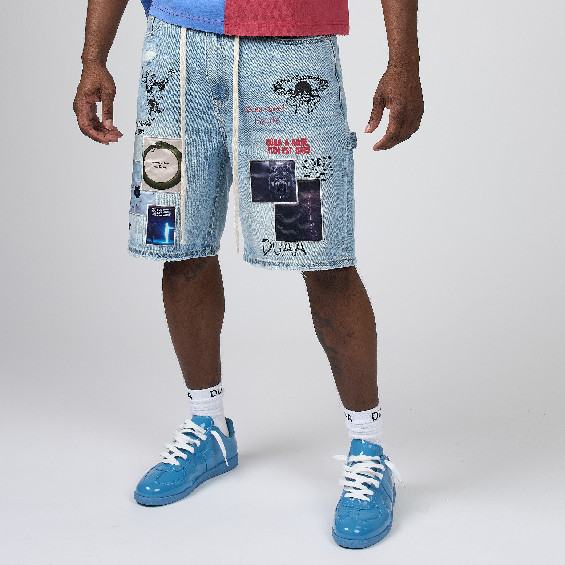 A person wearing light-wash denim shorts covered with graphic patches and stickers, paired with white socks and blue sneakers, photographed against a light gray studio background.