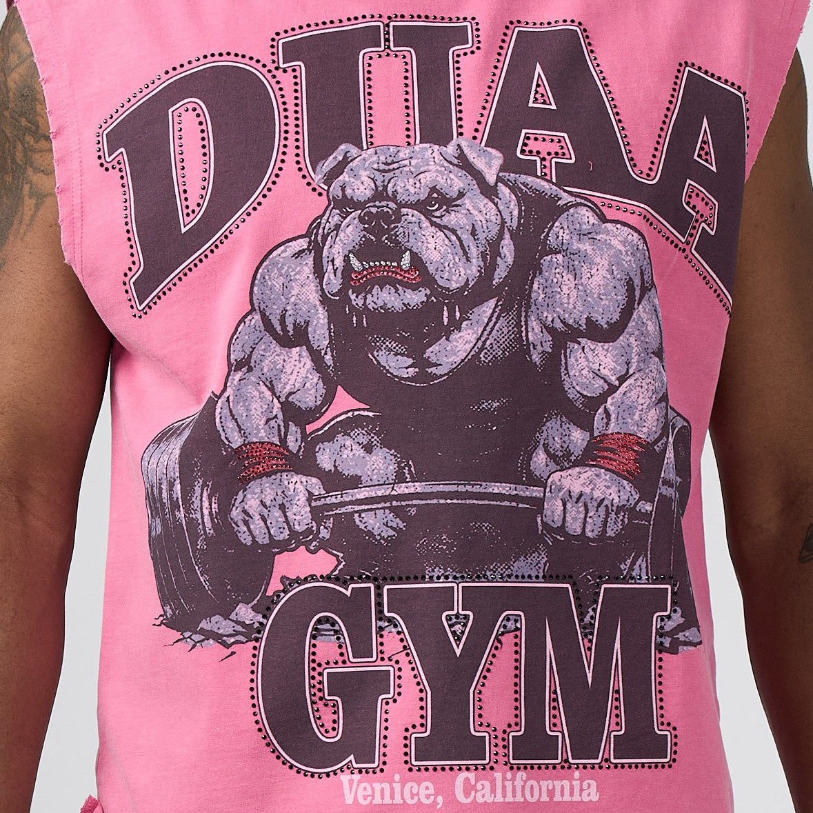 A man wearing a pink sleeveless muscle shirt with a graphic of a muscular bear and the text 'DUAA GYM' in dark brown, posed against a light gray studio background.